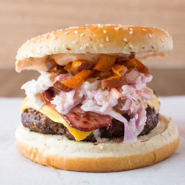 A Quickie's burger with cheese, bacon, coleslaw, and sweet potato fries on a sesame seed bun.