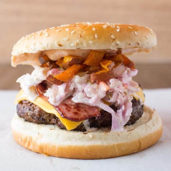 A Quickie's burger with cheese, bacon, coleslaw, and sweet potato fries on a sesame bun.