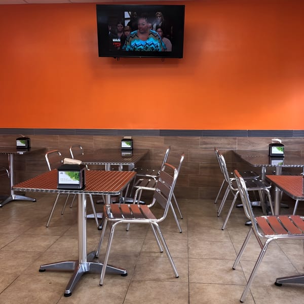 Empty dining area with orange walls, tiled floors, and metal tables and chairs, with a TV on the wall.