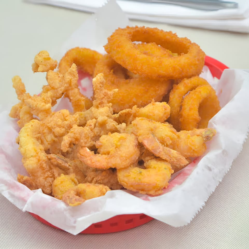 A red basket filled with golden-brown fried shrimp, clams, and onion rings from Quickie's Burgers, Wings & Seafood.