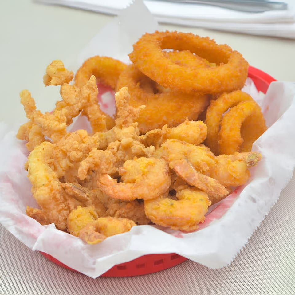 A red basket filled with golden-brown fried shrimp, clams, and onion rings from Quickie's Burgers, Wings & Seafood.