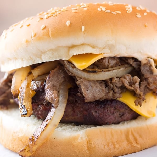 A Quickie's burger with a patty, melted cheese, grilled onions, and steak on a sesame seed bun.