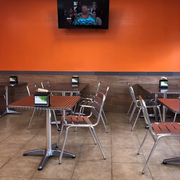 Empty dining area with orange walls, tiled floors, metal tables, and chairs, with a TV screen showing people.