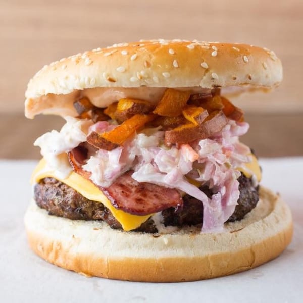 A Quickie's burger with cheese, bacon, coleslaw, and sweet potato fries on a sesame bun.
